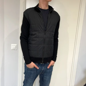 Svart quiltad cardigan från Belstaff - Snygg svart quiltad väst från Belstaff med dragkedja framtill. Västen har en stilren design med ribbade detaljer vid krage och ärmslut. Perfekt för lager-på-lager under kyligare dagar./ Aldirg använd