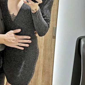 Svart glittrig klänning från Motel - Snygg svart glittrig klänning från Motel med djup v-ringning. Perfekt för en kväll ute! Klänningen har långa ärmar och en tight passform som framhäver figuren.