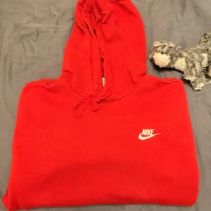 Röd hoodie från Nike - Säljer en snygg röd hoodie från Nike med klassisk logga på bröstet. Tröjan har en stor ficka framtill och justerbar huva med dragsko. Perfekt för en avslappnad stil.