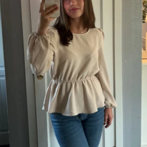 Beige blus med resår i midjan - Säljer en stilren beige blus med resår i midjan som ger en fin siluett. Blusen har långa ärmar med lätt puff och rund halsringning. Perfekt för en chic och avslappnad look.