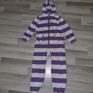 Lila och vit randig OnePiece för barn - Säljer en mysig lila och vit randig OnePiece för barn. Den har en dragkedja framtill och en praktisk huva. Perfekt för att hålla sig varm och bekväm hemma. 🟣⚪ den har ett litet hål på höger ben vid knät.