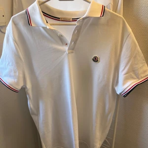 Vit pikétröja från Moncler - Säljer en stilren vit pikétröja från Moncler med korta ärmar och klassisk krage. Tröjan har snygga ränder i rött och svart på kragen och ärmsluten samt Monclers logga på bröstet. Perfekt för en avslappnad men ändå elegant look. Kvitto samt lappar från originalköp fås med!