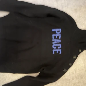 Svart tröja från Zadig & Voltaire med peace text  - Snygg svart tröja från Zadig & Voltaire med texten 'PEACE' i blått på framsidan. Tröjan har knappar längs axeln för en cool detalj. Perfekt för en stilren look. I ull använd två ggr 