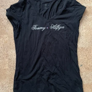 Svart t-shirt från Tommy Hilfiger - Snygg svart t-shirt från Tommy Hilfiger med v-ringning och korta ärmar. Perfekt för en stilren look med det ikoniska märket broderat på bröstet. En klassiker i garderoben!