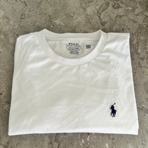 Vit t-shirt från Ralph Lauren - Säljer en klassisk vit t-shirt från Ralph Lauren med en broderad blå logotyp på bröstet. T-shirten har en slim fit passform och är tillverkad i mjuk bomull. Perfekt för en stilren look.