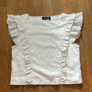 Vit topp från Pieces - Säljer en söt vit topp från Pieces med volangdetaljer på ärmarna. Toppen har ett diskret broderat mönster och är perfekt för en somrig look. Den är ärmlös och har en luftig passform. Aldrig använt.