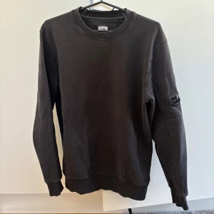 Svart tröja från C.P. Company - Säljer en stilren svart tröja från C.P. Company. Tröjan har långa ärmar och en rund halsringning. På ärmen finns en snygg detalj med C.P. Company-loggan. Perfekt för en casual look.