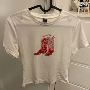 Vit t-shirt med cowboyboots från Shein - Säljer en vit t-shirt från Shein med ett tryck av röda cowboyboots och texten 'Austin Texas' på framsidan. Perfekt för en avslappnad stil med en touch av westernkänsla. T-shirten har korta ärmar och rund halsringning.