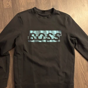 Svart tröja från BOSS - Snygg svart tröja från BOSS med ett stort logotryck i blått och svart på framsidan. Tröjan har långa ärmar och en rund halsringning. Hör av er vid minsta fråga. Nypris 1800 