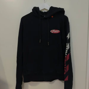 Svart hoodie från Off-White - Säljers en svart hoodie från Off-White med tryck i vitt och rött på ärmen och ryggen. Hoodien har en klassisk passform med dragsko i huvan och en stor ficka framtill. Perfekt för en stilren och modern look. Sannolik storlek L, nu cirka M.