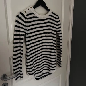 Randig stickad tröja från Gina Tricot - Säljer en snygg randig stickad tröja från Gina Tricot i svart och vit. Tröjan har långa ärmar och dekorativa knappar på axeln. Perfekt för en stilren look!