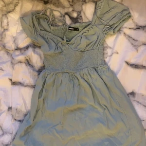 Ljusgrön klänning från BikBok - Charmig ljusgrön klänning från BikBok med smockdetaljer i midjan och korta puffärmar. Perfekt för en somrig look med sin mjuka och luftiga design. Klänningen har en söt v-ringning och är tillverkad i ett lätt material.