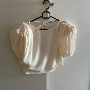 Beige blus från Adoore - Beige blus från Adoore med puffiga ärmar och rund hals. Blusen har en öppen rygg upptill. Storlek 38, axelvaddar