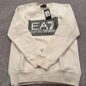 Vit sweatshirt från Emporio Armani - Säljer en stilren vit sweatshirt från Emporio Armani med ett coolt EA7-tryck i svart och silver på framsidan. Tröjan har långa ärmar och en klassisk rund halsringning. Perfekt för en avslappnad och trendig look.