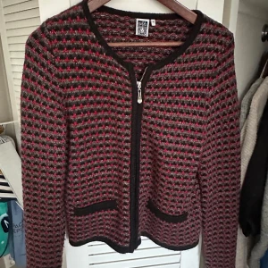 Mönstrad kofta från Isolde - Säljer en snygg kofta från Isolde med ett unikt mönster i rött och grått. Koftan har en dragkedja framtill och två praktiska fickor. Perfekt för att hålla sig varm och stilren under kyligare dagar.