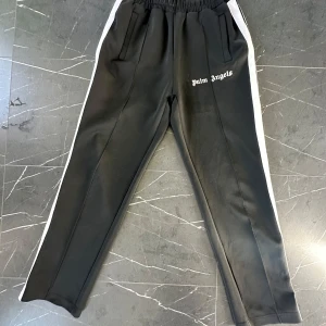 Palm Angels byxor - Snygga svarta träningsbyxor från Palm Angels med vita sidoremsor och elastisk midja. Byxorna har en avslappnad passform och är perfekta för en sportig look. Tillverkade i polyester för en bekväm känsla.