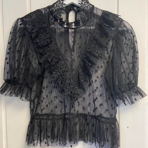 Svart spetsblus med volangdetaljer - Elegant svart blus i spets med volangdetaljer och prickigt mönster. Blusen har trekvartsärmar och en hög hals med knäppning i nacken. Perfekt för en stilfull look😍✨