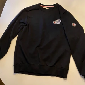 Svart tröja från Moncler storlek S - Säljer en stilren svart tröja från Moncler med broderade logotyper på bröstet och ärmen. Tröjan har en klassisk passform och rund halsringning. Perfekt för en avslappnad och trendig look.