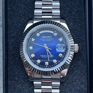 Seiko day date  - Snygg silverfärgad klocka från seiko med en blå urtavla och dag- och datumvisning. Urtavlan har glittrande stenar vid varje timme och en räfflad bezel. Armbandet är i metall med en klassisk länkdesign.