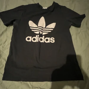 Svart t-shirt från Adidas - Säljer en svart t-shirt från Adidas med det klassiska trefoil-loggan i vitt på framsidan. T-shirten är kortärmad och har en normal passform. Perfekt för en sportig look eller avslappnad stil.