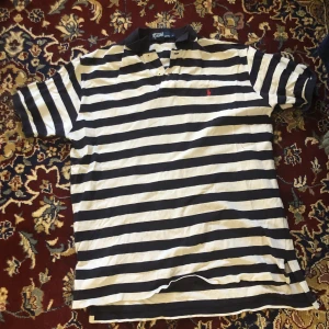 Randig pikétröja från Ralph Lauren - Säljer en klassisk svartvit randig pikétröja från Ralph Lauren. Tröjan har korta ärmar och en liten röd logga på bröstet. Perfekt för en stilren look.