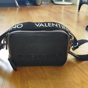 Svart axelväska från Valentino - Snygg svart axelväska från Valentino med dubbla dragkedjor och justerbar axelrem. Väskan har en stilren design med logotypen tryckt på framsidan och axelremmen. Perfekt för att bära dina nödvändigheter med stil. Använd 3 gånger. Nypris 1900:-