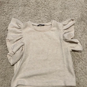 Beige topp från Zara med volangärmar - Säljer en beige topp från Zara med volangärmar. Toppen har en normal passform och är tillverkad i ett mjukt material. 