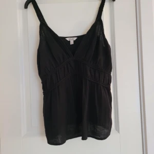 Svart linnetopp från Aware - Snygg svart linnetopp från Aware med v-ringad design och flätade detaljer. Perfekt för varma dagar eller som ett lager under en kavaj. Den har smala axelband och en luftig passform.