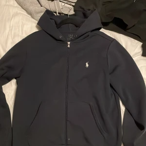 Mörkblå hoodie från Ralph Lauren - Säljer en stilren mörkblå hoodie från Ralph Lauren med dragkedja och det klassiska broderade logotypen på bröstet. Perfekt för en avslappnad look. Den har en bekväm passform och är tillverkad i mjukt material.