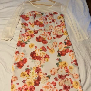 Blommig klänning med trekvartsärm😄 - Charmig klänning med blommigt mönster i rött, gult och grönt på en vit bas. Den har trekvartsärmar och en elegant passform som passar perfekt för vår och sommar.