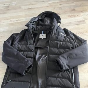 Svart pufferjacka från Columbia - Säljer en svart pufferjacka från Columbia med huva och dragkedja. Jackan har långa ärmar och är perfekt för kyligare väder. Den har en stilren design med Columbia-loggan på bröstet och ärmen.