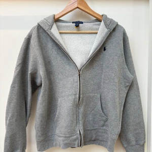 Grå hoodie med dragkedja från Polo Ralph Lauren - Grå hoodie från Polo Ralph Lauren med dragkedja och två fickor framtill. Mjuk insida och klassisk broderad logga på bröstet. Perfekt för en avslappnad stil och enkel att matcha med det mesta.