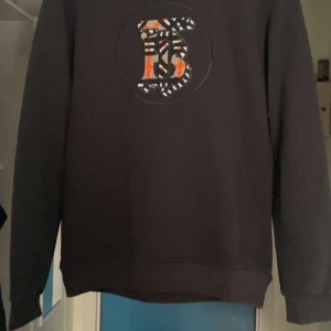 Svart sweatshirt från Burberry med broderad logga - Säljer en svart sweatshirt från Burberry i 100% bomull. Tröjan har en stor broderad Burberry-logga i vitt och orange framtill. Klassisk passform med rund hals och långa ärmar. Perfekt för en stilren och trendig look.
