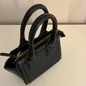 Svart handväska med kedjerem - Säljer en stilren svart handväska med krokopräglat mönster och guldfärgad dragkedja. Väskan har både handtag och en avtagbar axelrem i kedja och skinn. Perfekt för dig som vill ha en trendig accessoar till din outfit.