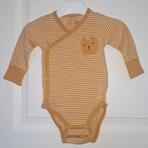 Randig omlottbody med björnficka från Lindex - Supergullig omlottbody från Lindex i senapsgult och vitt med smala ränder. En söt liten ficka framtill i form av ett björnhuvud. Extra långa ärmar & dubbel knäppning, för extra långbarhet. Perfekt för de minsta att växa i!