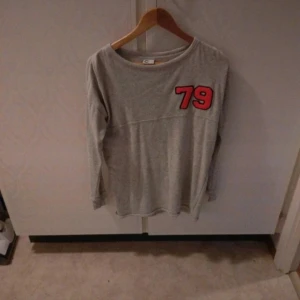 Grå långärmad tröja med röd 79-dekal - Säljer en grå långärmad tröja från C i storlek XS. Tröjan har en röd och svart 79-dekal på bröstet och är tillverkad i mjukt bomullsmaterial. Perfekt för en avslappnad stil.