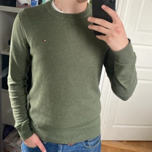 Olivgrön tröja från Hilfiger Denim - Säljer en olivgrön tröja från Hilfiger Denim med rund halsringning och diskret logga på bröstet. Tröjan har långa ärmar och ribbade muddar vid ärmslut och nederkant. Perfekt för en stilren och avslappnad look.  Sömmen på storleks etiketten har lossnar men syns ej vid användning.