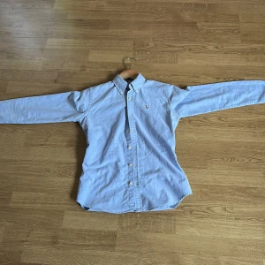 Ljusblå skjorta från Ralph Lauren - Säljer en klassisk Helt ny oanvänd  ljusblå skjorta från Ralph Lauren med button-down krage och broderad logga på bröstet. Skjortan har långa ärmar och knäppning framtill. Perfekt för sommaren.