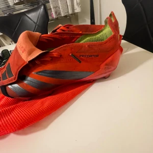Röd orange fotbollsskor från Adidas - Säljer ett par röda Adidas Predator fotbollsskor med svarta detaljer. Skorna har snörning och är designade för optimal prestanda på planen. Perfekta för fotbollsspelare som vill ha både stil och funktion. Väskan ingår.