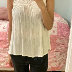 Vit ärmlös blus med spets från KappAhl - Säljer en söt vit blus från KappAhl med spets upptill och plisserat tyg nedtill. Blusen är ärmlös och har en rund halsringning. Perfekt för varma dagar eller sommarens alla tillfällen.