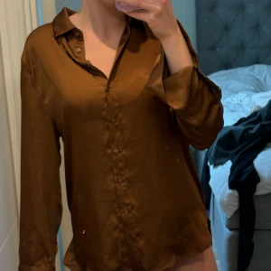Brun satinblus från Gina Tricot - Säljer en brun, glansig satinblus från Gina Tricot. Skjortan har klassisk krage, knappar framtill och långa ärmar. Perfekt för dig som gillar en stilren och trendig look.