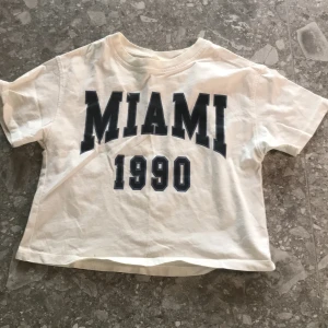 Vit croppad t-shirt med MIAMI-tryck - Säljer en vit croppad t-shirt från KappAhl med stort svart MIAMI 1990-tryck på framsidan. T-shirten har korta ärmar och rund halsringning. Perfekt för en avslappnad och cool stil.