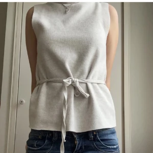 Ljusgrå ärmlös topp med knytband och hög krage  - Säljer en stilren beige ärmlös topp med knytband i midjan. Toppen har en rak passform och är tillverkad i ett mjukt material som passar perfekt till jeans eller kjol. Hög krage 