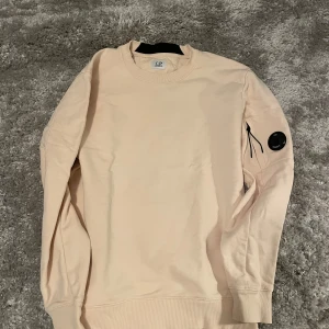 Beige tröja från C.P. Company - Säljer en stilren beige tröja från C.P. Company. Tröjan har långa ärmar och en unik detalj med en rund svart logotyp på ärmen. Perfekt för en avslappnad och trendig look. Aldrig använd