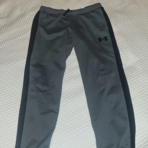 Under Armour träningsbyxor  - Grå träningsbyxor från Under Armour med elastisk midja och dragsko. Byxorna har en diskret logga på ena benet och är perfekta för en avslappnad stil. Junior XL passar som S herr.