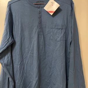 Blå långärmad tröja från Jockey - Säljer en blå långärmad tröja från Jockey med rund halsringning och knappslå framtill. Tröjan har en bröstficka och är tillverkad i mjuk bomull. Perfekt för en avslappnad stil.