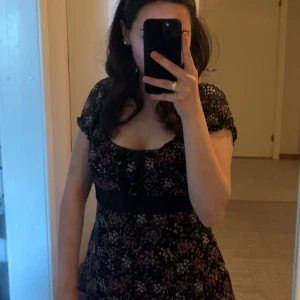 Blommig blus med knytband - Charmig svart blus med blommigt mönster i rosa och vitt. Blusen har korta ärmar och ett knytband i midjan för en smickrande passform. Perfekt för en somrig look! Pris kan diskuteras🫶🏼