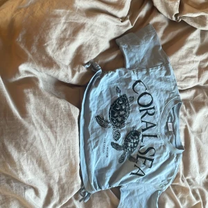 Blå t-shirt med sköldpaddor från Lindex - Säljer en blå magtröja från Lindex med tryck av sköldpaddor och texten 'Coral Sea'. T-shirten har korta ärmar och en knytning i nederkant. Perfekt för en avslappnad stil!