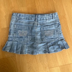 Jeanskjol med volang och broderade fickor - Säljer en ljusblå jeanskjol med volangkant och broderade kronor på bakfickorna. Kjolen har fickor både fram och bak samt bälteshällor. Perfekt för dig som gillar en cool och avslappnad stil.