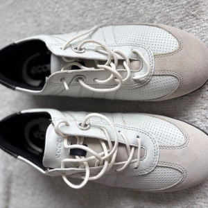 Vita sneakers från Ecco med svarta detaljer - Säljer ett par vita sneakers från Ecco med svarta detaljer och snörning. Skorna har ovandel i skinn och mocka, perforerade partier och rund tå. Snygg och sportig design som passar till många outfits.